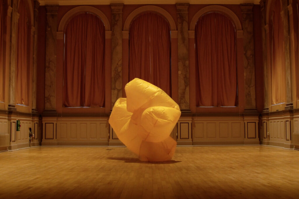Bild från performanceverket cmyk. En dansare rör sig inne i en stor gul uppblåst ballongliknande dräkt.