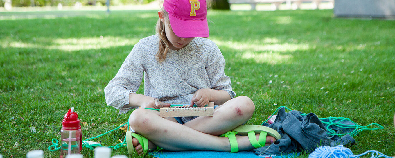 Ett barn sitter i gröngräset i Skulpturparken. I förgrunden färgtuber och annat material för kreativt skapande.