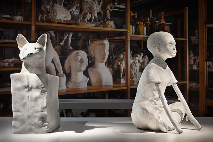 Two plaster sculptures on a table. One depicts a fox sticking its head out of a bag or box. The other depicts a boy sitting in a tailor's pose with his eyes closed. His arms are made of tree branches.