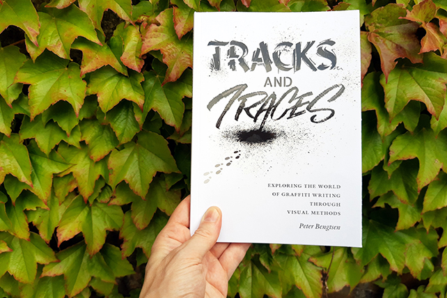 En hand håller upp boken "Tracks and Traces" av Peter Bengtsen. I bakgrunden växtlighet mot en vägg.
