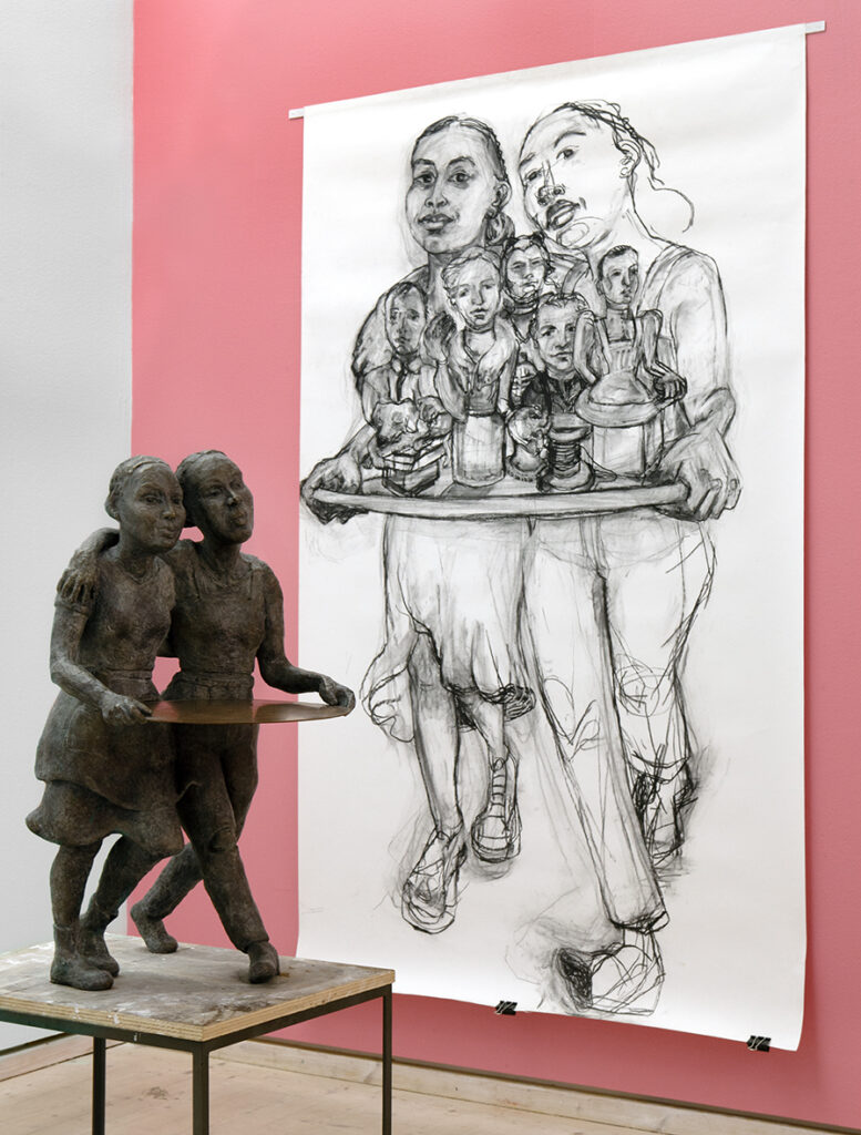 En bronsskulptur på ett podium och en stor skiss till samma skulptur hänger på väggen bakom. Väggen är rosa.