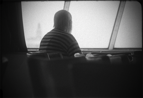 Svartvit stillbild från video. Ombord på en en färja. En man sitter och tittar ut genom ett fönster.