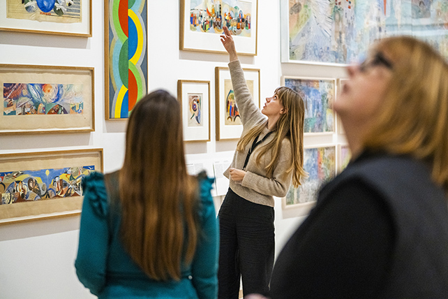 tre människor i bild. En museiguide visar färgstarka målningar på en vägg för två besökare.