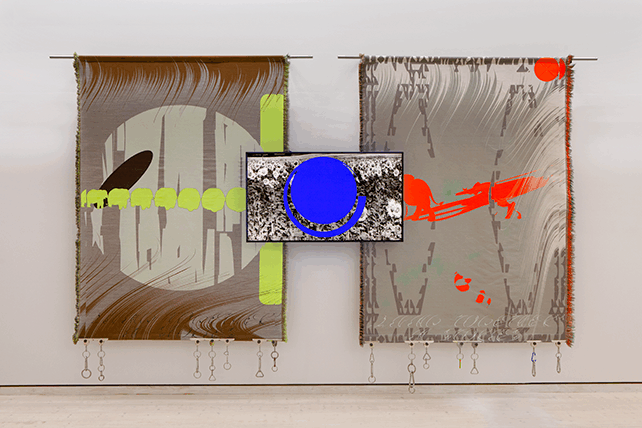 Två stora färgstarka vävar hänger mot en vit vägg. I mitten visas en video på skärm.