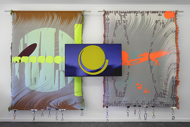 Två stora färgstarka vävar hänger mot en vit vägg. I mitten visas en video på skärm.