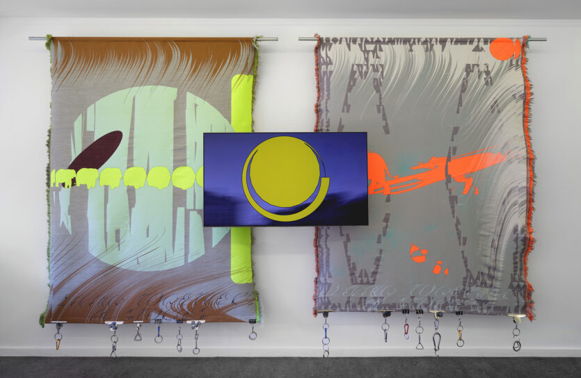 Två stora färgstarka vävar hänger mot en vit vägg. I mitten visas en video på skärm.