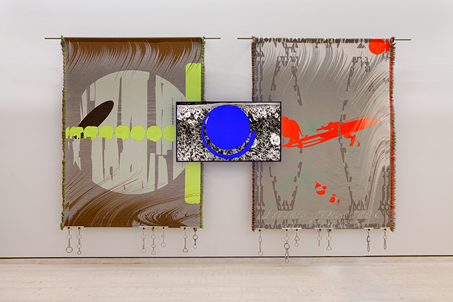 Två stora färgstarka vävar hänger mot en vit vägg. I mitten visas en video på digital skärm.