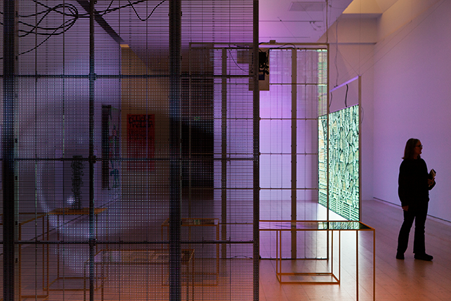 Stor transparent bildskärm visar ett öga, lila ljus. I bakgrunden en annan skärm med grönt ljus, en människa tittar på något utanför bilden.