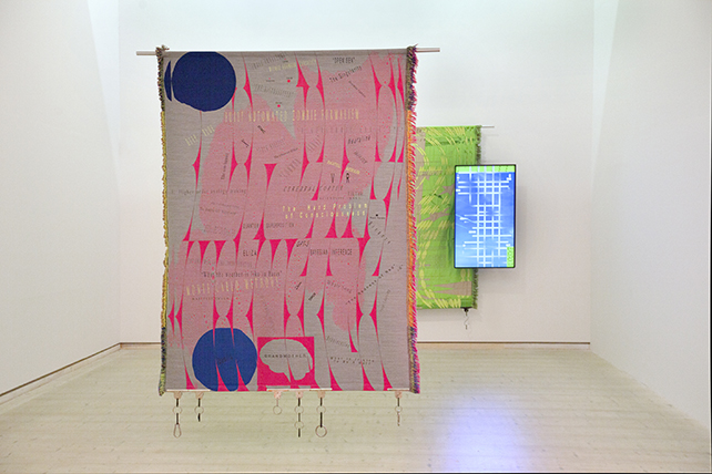 En konstväv i rosa, grått och blått. I bakgrunden en annan väv i grönt tillsammans med en videoskärm.