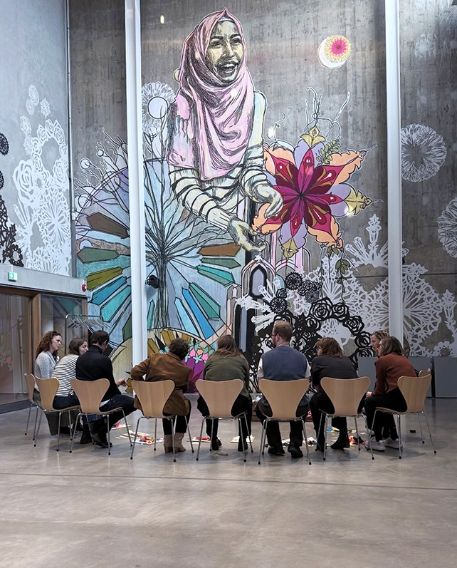 Människor sitter i en halvcirkel och betraktar ett stort väggkonstverk.