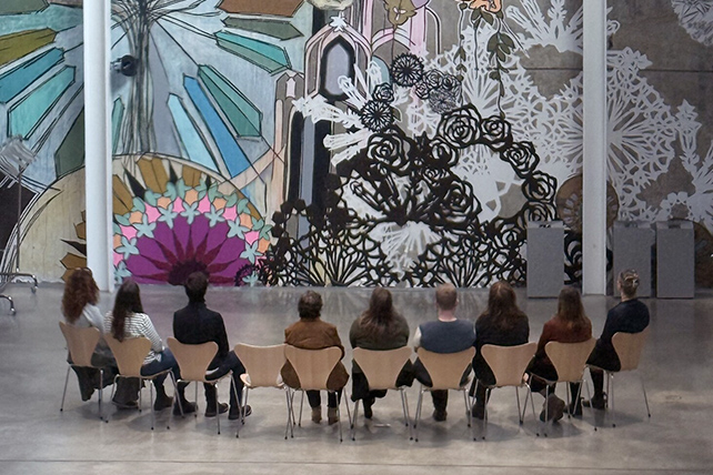 Människor sitter i en halvcirkel och betraktar ett stort väggkonstverk.