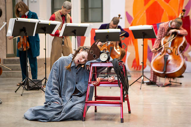 En skådespelare sitter på golvet med huvudet lutat mot en pall. Hon gäspar. I bakgrunden musiker.